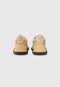 Zapatillas beige con parte superior de malla y gamuza, que presentan un talón texturizado y una suela de goma oscura. Dos tiradores en la parte posterior.