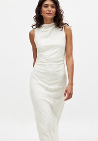 Valkoinen hihaton mekko, jossa on korkea kaula-aukko, ja jossa on teksturoitu kukkakuosi sekä rypytys vyötäröllä, valmistettu sileästä kankaasta.