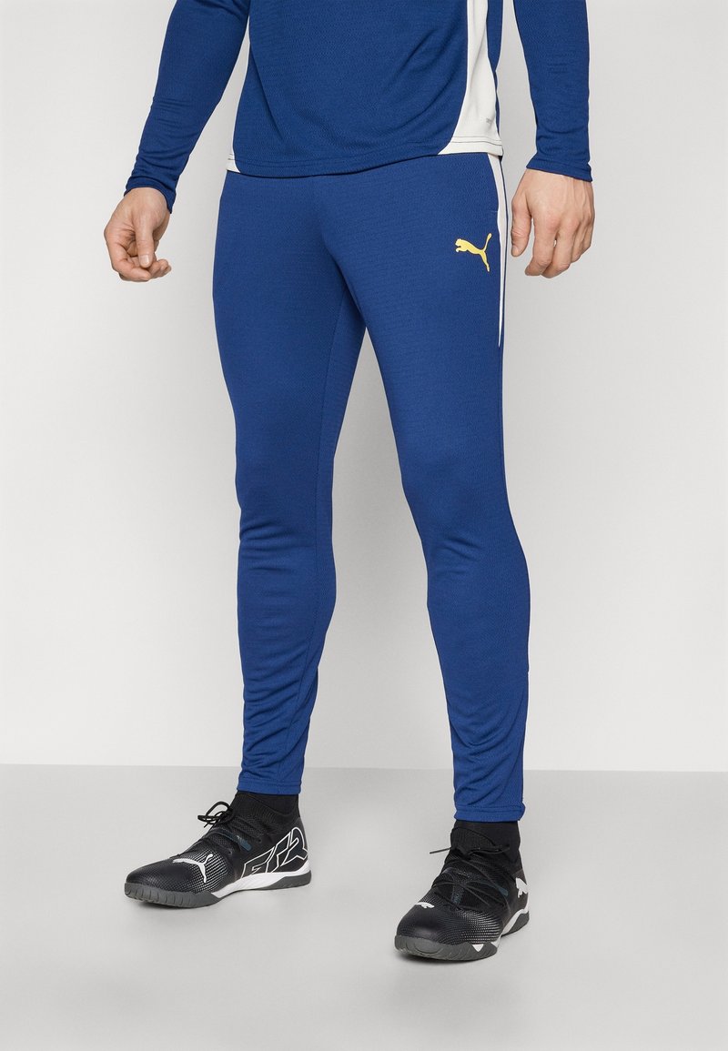 Pantalon de sport bleu en matériau lisse, avec une coupe ajustée, une taille élastique, et un petit logo jaune sur la cuisse gauche.
