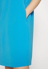 Modré šaty s hladkou texturou, vybavené bočními kapsami a rovnou siluetou. Tkanina vypadá lehce a sahá po kolena.