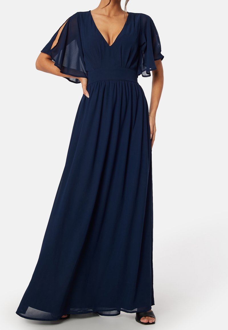 Femme portant une robe longue bleu marine avec un décolleté en V profond et des manches courtes fluides avec des fentes, debout avec une main sur la hanche.