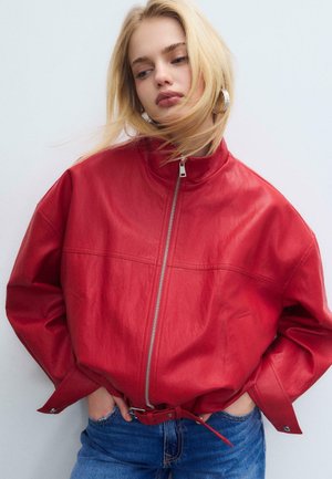 Mladá žena se světlými vlasy, která nosí stříbrné kruhové náušnice, červenou koženou bundu na zip a modré džíny, stojící s rukama v kapse.