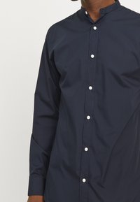 Mörkblå skjorta med mandarin krage, tillverkad av glatt tyg, med vita knappar längst fram och långa ärmar.