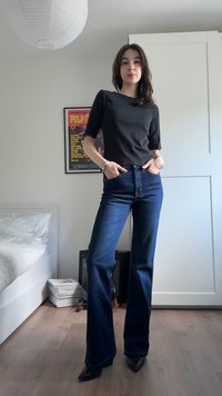 Tânără în top negru și blugi albastru închis stă cu încredere într-un dormitor modern cu echipament muzical și un poster încadrat.