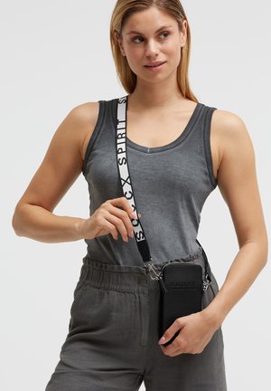 Woman wearing gray sleeveless top and pants, holding black crossbody phone pouch with white strap labeled "SPIRIT."
