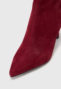 Stivaletto alla caviglia in suede rosso con punta affusolata e tacco a stiletto. La parte superiore ha una texture liscia e dettagli di cucitura minimi.