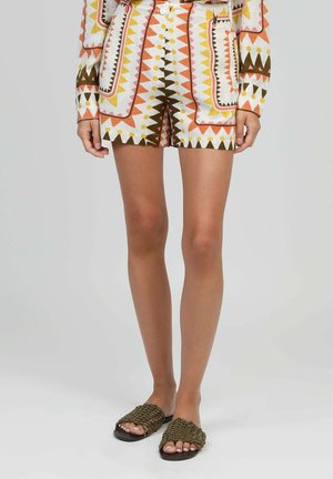 Jambes portant un short à motifs et une veste assortie avec des formes géométriques en orange, jaune, marron et blanc, associés à des sandales marron tissées à enfiler.