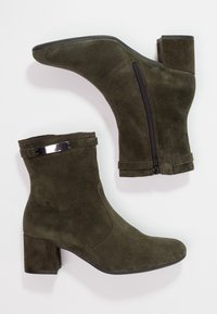 Botines de ante en verde oliva oscuro con un tacón cuadrado grueso, cremallera lateral y una banda de acento plateado en el puño. Textura suave.