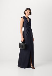 Navy satin maksimekko, jossa on solmittu etuosasuunnittelu ja korkea halkio. Malli pitää pientä mustaa käsilaukkua ja hänellä on jalassaan mustat rannerenkaat.