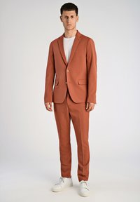 Conjunto de color óxido con un blazer de un solo botón y pantalones a juego. Camiseta blanca por debajo, combinada con zapatillas blancas. Textura suave.