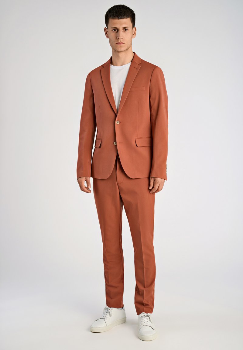 Conjunto de color óxido con un blazer de un solo botón y pantalones a juego. Camiseta blanca por debajo, combinada con zapatillas blancas. Textura suave.