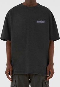 Camiseta de algodón negra oversize con mangas cortas, que presenta un pequeño texto gráfico morado en el pecho. Textura suave y diseño casual.