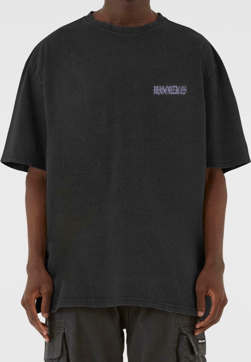Camiseta de algodón negra oversize con mangas cortas, que presenta un pequeño texto gráfico morado en el pecho. Textura suave y diseño casual.