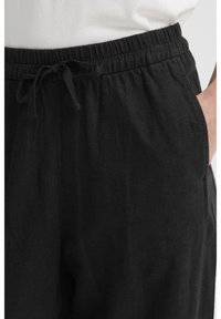 Μαύρο λινό παντελόνι με κορδόνι και ελαστική μέση, φορεμένο με το ένα χέρι στην τσέπη και λευκό πουκάμισο κρυμμένο μέσα.