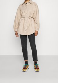 Persona che indossa un cappotto beige con cintura, jeans neri tagliati e sneakers colorate con brillanti lacci blu, in piedi davanti a uno sfondo semplice.