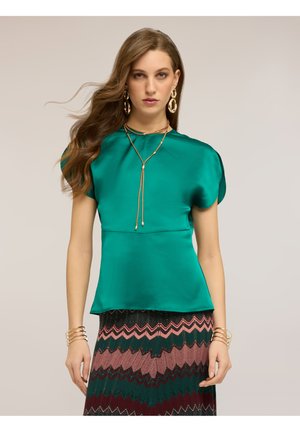 Kobieta w turkusowej satynowej bluzce, wzorzystej spódnicy z motywem zygzaków, złotym, warstwowym naszyjniku, dużych kolczykach i wielu złotych bransoletkach stoi na jednolitym tle.