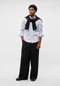 Ανοιχτό μπλε πουκάμισο με κουμπιά, μαύρο πουλόβερ δεμένο στους ώμους, μαύρο παντελόνι με φαρδιά μπατζάκια και μαύρα χοντρά παπούτσια. Απλό φόντο.