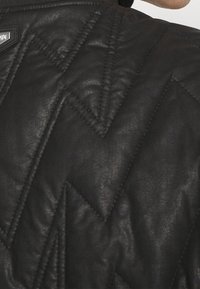 Veste matelassée noire avec un motif en chevrons diagonal, en matériau synthétique lisse avec une finition texturée et un logo de marque.