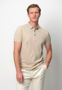 Beige pikétröja med korta ärmar, tvåknapplås och mörka knappar; minimal textur och en avslappnad silhuett. Buren med ljusa byxor.