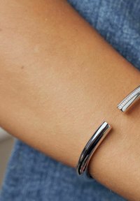 Silverfärgad armband i form av en manschett med en slät, polerad yta, som har en elegant design och en detalj av små inlagda kristaller på ena sidan.
