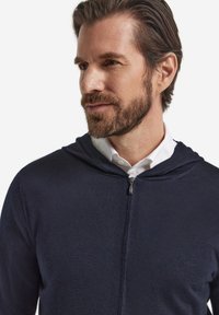 Mörkblå zip-hoodie i mjukt tyg, med huva, silverfärgad dragkedja och strukturerad yta, bärs över en vit skjorta med krage.