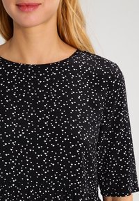 Top nero con scollo a girocollo, caratterizzato da un motivo di piccoli cuori bianchi e una finitura a peplum. Il materiale sembra leggero e liscio.