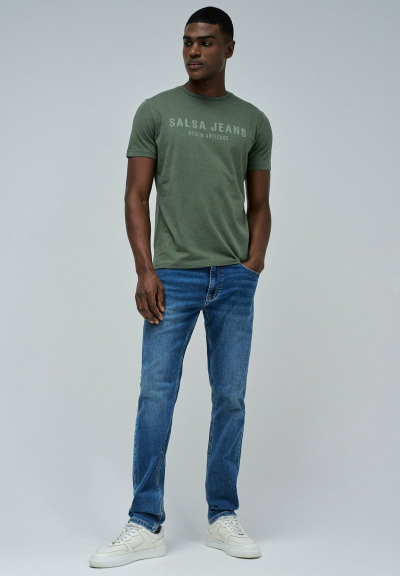 Camiseta verde de manga corta con "SALSA JEANS" impreso, combinada con unos vaqueros de mezclilla azules y zapatillas blancas. Fondo liso.