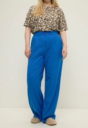 Person wearing a leopard print short-sleeve top, bright blue high-waisted wide-leg pants, and beige fuzzy slippers standing against plain background.