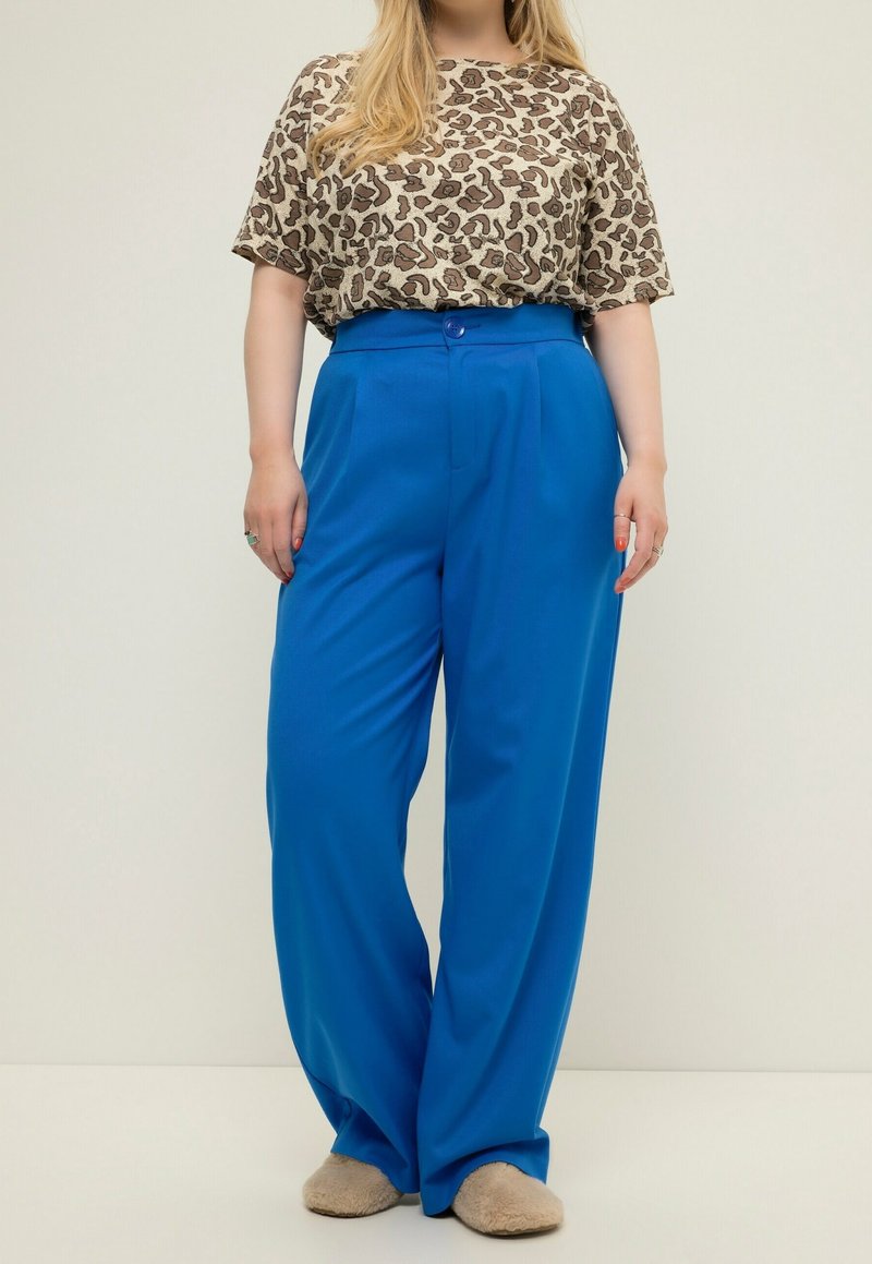 Personne portant un haut à manches courtes imprimé léopard, un pantalon large taille haute bleu vif et des chaussons beiges duveteux, debout devant un fond uni.