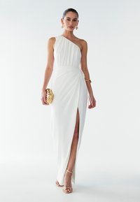 Vit, asymmetrisk klänning med draperad överdel, lårhög slits och knytning i midjan, matchad med guldfärgad clutcht och remsandaler.