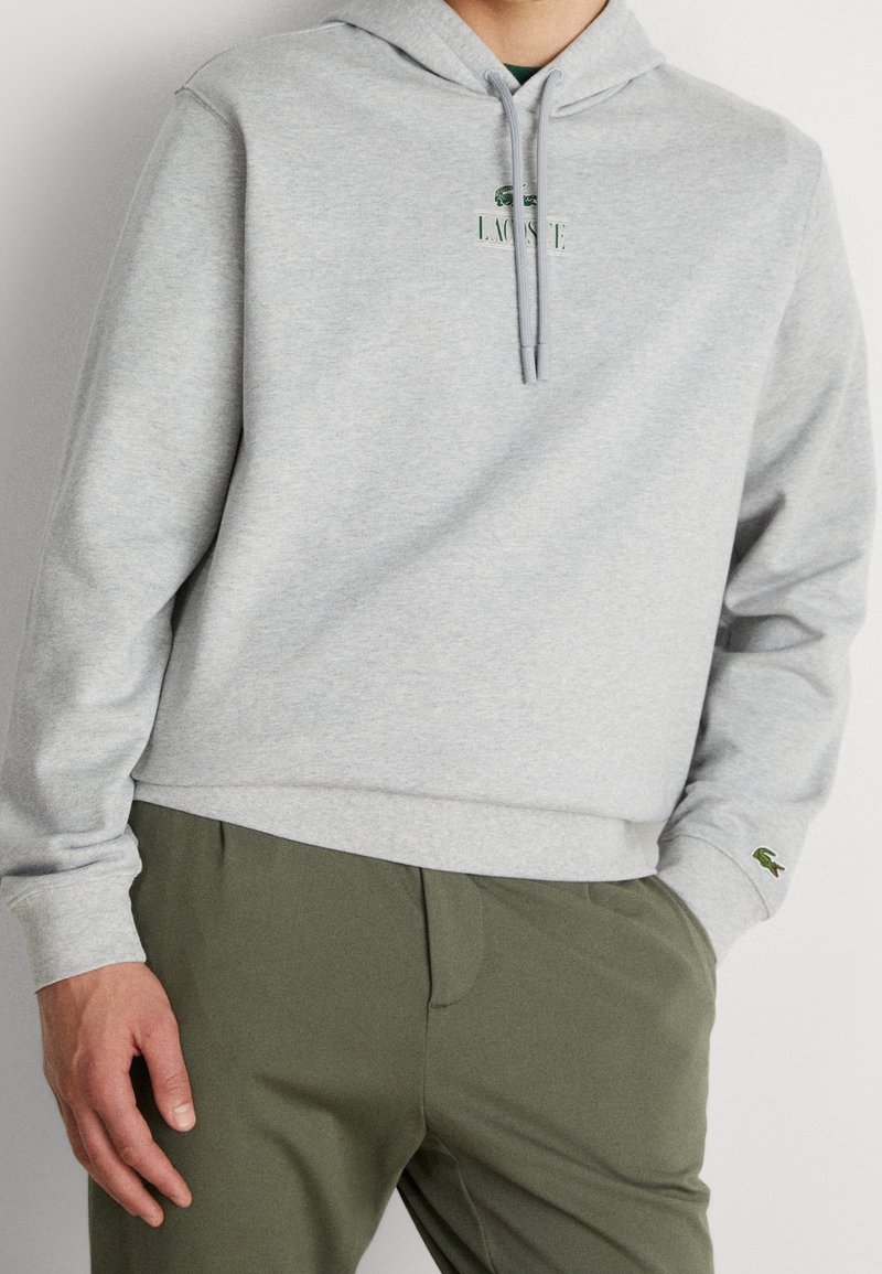 Homme portant un sweat à capuche Lacoste gris clair avec cordons et pantalon vert olive, main reposant sur le côté, vue partielle de face.