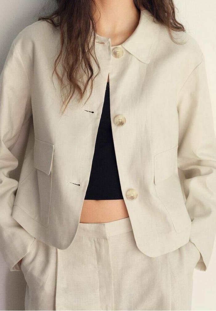 Femme portant une veste beige courte boutonnée et un pantalon taille haute assorti avec un top court noir, mains dans les poches, cheveux lâchés sur les épaules.