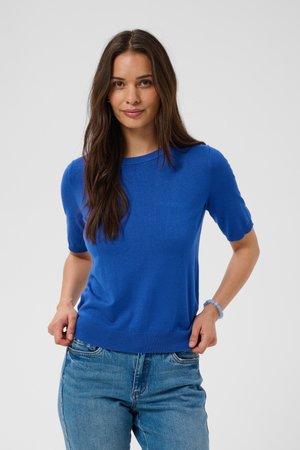Femme aux longs cheveux bruns portant un pull bleu vif à manches courtes et un jean bleu clair, debout avec les mains près des poches.