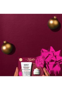 Hands holding Kiehl's Ultra Facial Cleanser and Heavy Eye Treatment against a maroon background with two gold ornaments and a pink bow.