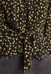 Czarna bluzka z tkaniny ozdobiona żółtym wzorem kwiatowym, z wiązanym przodem i subtelnymi falbanami u dołu, zestawiona z szarymi dżinsami.