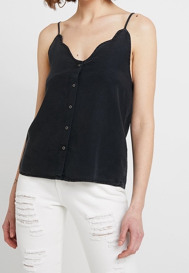 Femme portant un caraco noir boutonné à l'avant avec un décolleté festonné et un jean skinny blanc déchiré, sur un fond uni.