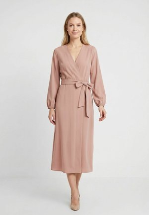 Femme portant une robe portefeuille à manches longues rose poudré avec un nœud noué à la taille, debout devant un fond clair uni.