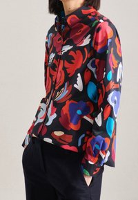 Blouse à manches longues en tissu noir avec un motif floral vibrant en rouge, bleu et vert. Comprend un col à boutons et des poignets.