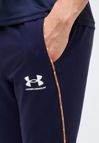 Χέρι μερικώς στην τσέπη του σκούρου μπλε παντελονιού Under Armour με πορτοκαλί περιμετρική διακόσμηση και ορατό λογότυπο στον μηρό.