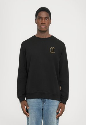 Zwarte sweatshirt met een geribde ronde hals, voorzien van een gouden geborduurd logo op de borst. Draagt met lichtblauwe jeans.