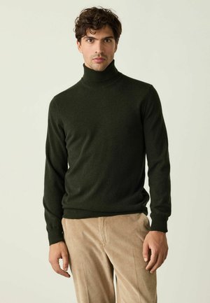 Junger Mann mit kurzen lockigen Haaren, der einen dunkelgrünen Rollkragenpullover und hellbraune Cordhosen trägt, steht vor einem einfarbigen Hintergrund.