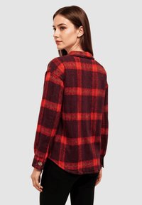 Camisa de franela a cuadros en rojo y negro con cuello, mangas largas, puños de botón y un dobladillo ligeramente curvado. Tejido texturizado con patrones audaces.