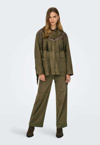 Giacca in suede verde oliva con dettagli frangiati, yoke in pelle marrone, grandi tasche anteriori, abbinata a pantaloni a gamba larga coordinati e stivali scuri.
