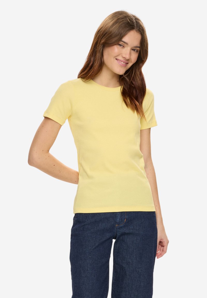 Femme aux longs cheveux bruns portant un t-shirt jaune ajusté à manches courtes et un jean bleu foncé, souriant avec une main derrière le dos.
