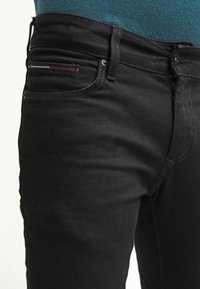 Černé džíny z denimu s hladkým povrchem, přední kapsa s červenými, bílými a modrými švy a standardní poutka na opasek.