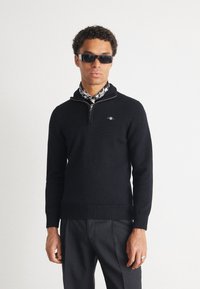 Pulover negru din tricot, cu guler cu jumătate de fermoar, tiv originat, și un mic emblemă pe piept, asortat cu ochelari de soare întunecați și o eșarfă cu imprimeu.