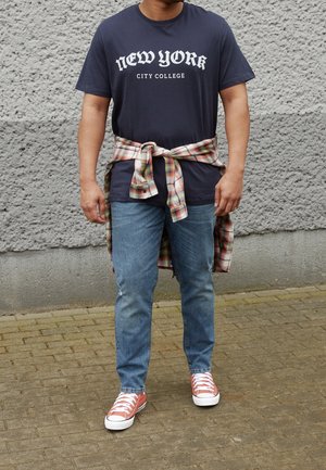 Camiseta de marinero con el texto "NUEVA YORK CITY COLLEGE", camisa de cuadros atada a la cintura, jeans azules y zapatillas rosas con suelas blancas.