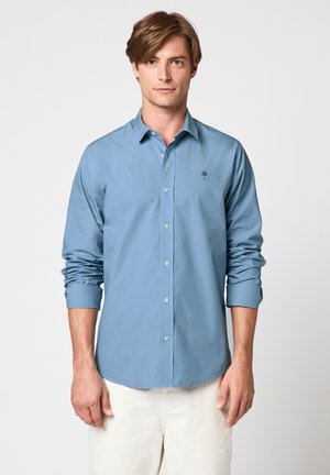 Jeune homme portant une chemise à manches longues bleu clair avec boutons et un pantalon blanc, debout devant un fond blanc uni.