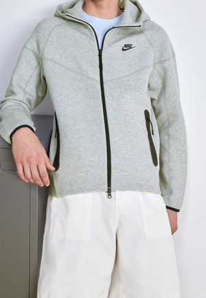 Person trägt einen hellgrauen Nike Zip-Hoodie mit schwarzen Reißverschlussdetails und weißen Hosen, legt die Hand auf einen Metallschrank.