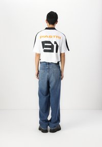 White short-sleeve shirt with black accents and large black "31" and "PIASTRI" text on the back, paired with loose blue denim jeans and black shoes.